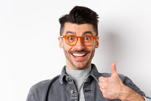 Close up portrait of excited funny guy in eyewear showing thumbs up, smiling supportive, praise and recommend cool store, standing on white background.