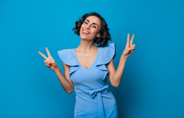 Close up portrait of an excited happy gorgeous girl in a long blue dress who is looking in the camera while she posing with a bright toothy smile and shows victory sign on the camera