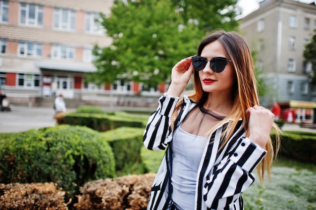 Close up portrait of fashionable woman look with black and white striped suit jacket and sunglasses posing against bushes at street. Concept of fashion girl.