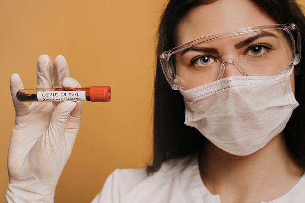 Close up portrait of a female doctor in surgical mask, gloves and protective glasses holds a test tube with a coronavirus Covid-19 test.Doctors, infectionist, research and covid19 concept.