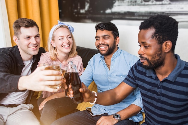 Photo close up portrait of four joyful friends clinking their glasses and bottles of drinks, smiling sitting on sofa at home. home party, celebration, people and holidays concept.