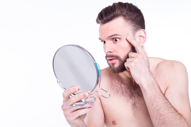 Close up portrait of frustrated naked bearded man looking in the mirror for acne on his face isolated on white background