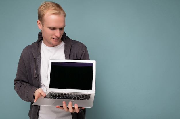 Close-up portrait of handsome blonde man holding computer laptop looking down at netbook keyboard isolated over blue background.