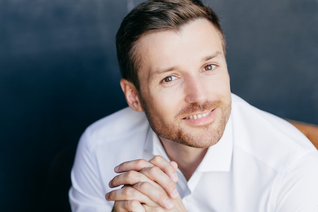 Close up portrait of handsome unshaven male with positive smile, keeps hands together