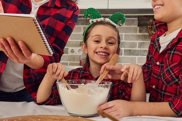 Close-up portrait of a happy cheerful sweet Caucasian baby girl smiles toothy smile looking at camera sitting at a kitchen table between her brother and mom while cooking together Christmas pastries