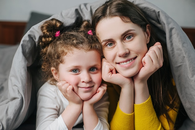 Close up portrait of happy cute little child kid daughter and cheerful young mother lying on bed covered with blanket look at camera posing at home, family mom and small girl spend time together