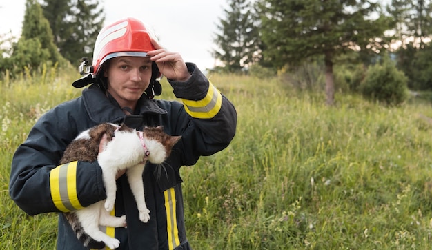 Close-up portrait of heroic fireman in protective suit and red helmet holds saved cat in his arms. Firefighter in fire fighting operation. High quality photo