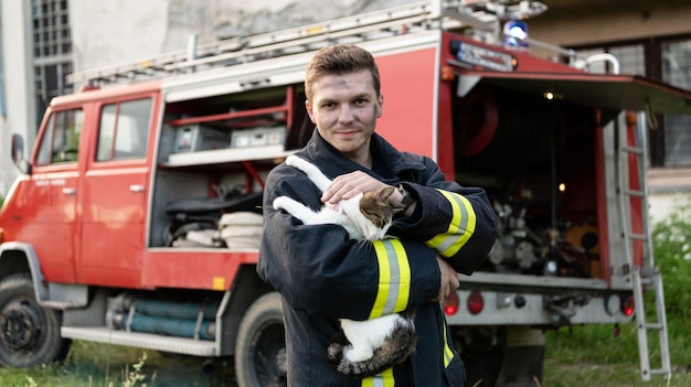 Close-up portrait of heroic fireman in protective suit and red helmet holds saved cat in his arms. Firefighter in fire fighting operation. High quality photo