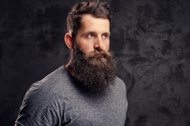 Close-up portrait of a hipster with full beard and stylish haircut, dressed in a gray t-shirt, stands in a studio on a dark background.