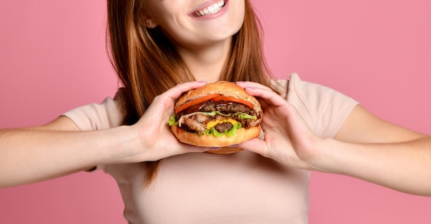 Close up portrait of a hungry young woman eating burger isolated over pink background