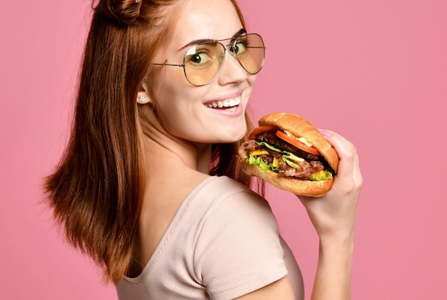 Close up portrait of a hungry young woman eating burger isolated over pink background