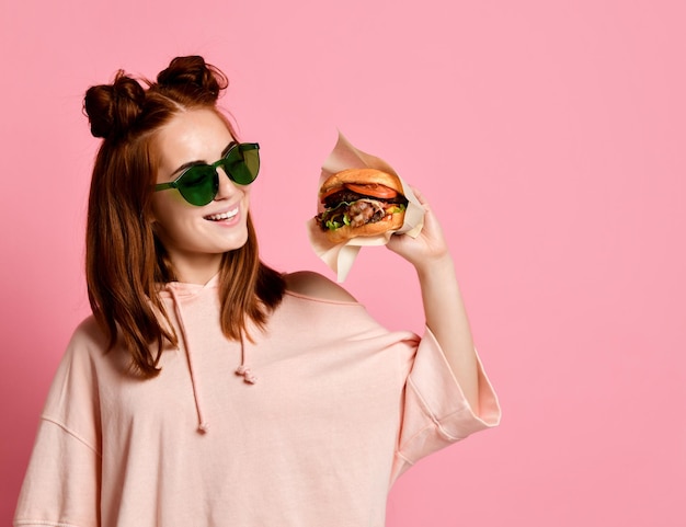 Close up portrait of a hungry young woman eating burger isolated over pink background