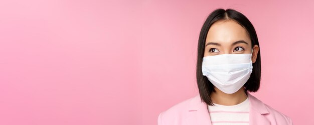 Close up portrait of japanese corporate woman in medical face mask from covid19 looking left at logo sale promo standing over pink background