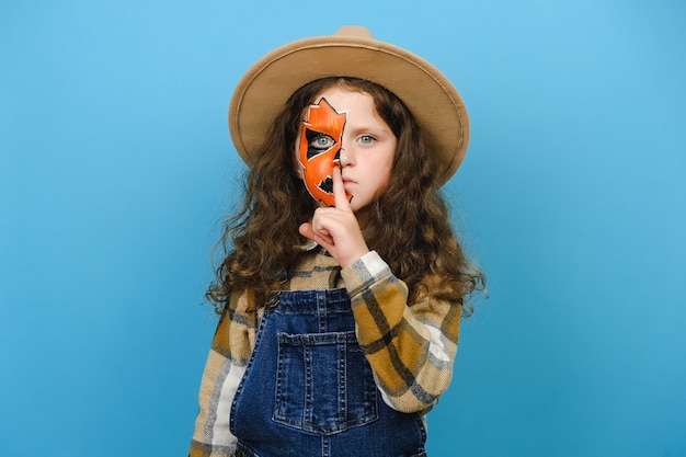 Close up portrait of little girl child with Halloween makeup mask wear hat and shirt, say hush be quiet finger on lip shhh gesture isolated on blue background studio. Celebration holiday party concept