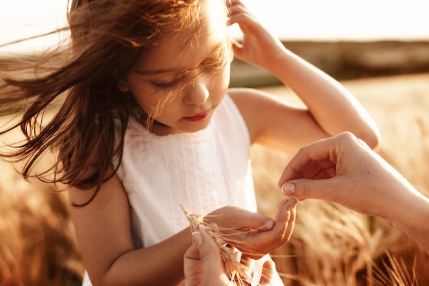 Close up portrait of a lovely little girl dressed in white dress holding wheat in hand while wind is playing with her hair against sunset.