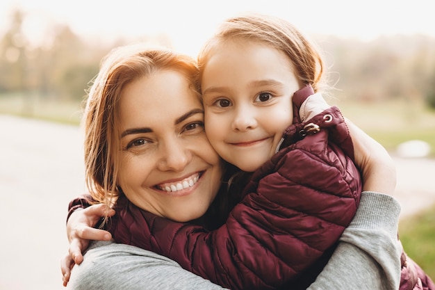 Close up portrait of a lovely mother embracing her daughter looking at camera laughing against sunset outdoor in the park.