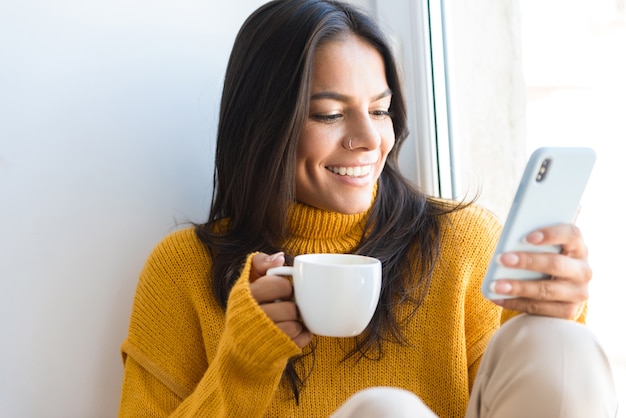 Close up portrait of a lovely young woman dressed in sweater sitting at the window indoors, holding cup of tea, using mobile phone