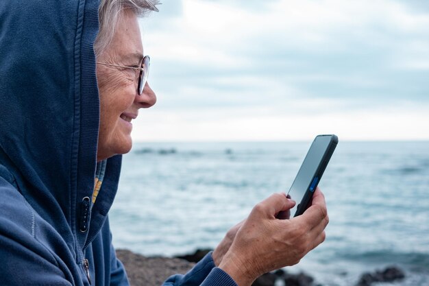 Close up portrait of mature senior woman enjoying retirement at sea Caucasian elderly female standing on the beach in a cloudy day using mobile phone enjoying tech and social Horizon over water