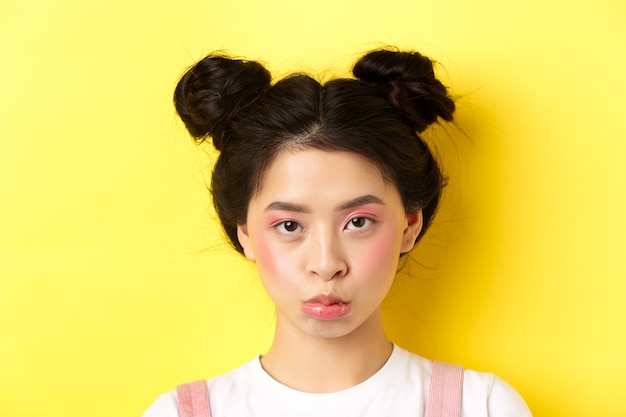 Close up portrait of moody asian girl pouting and looking upset at camera, standing wiht glamour makeup and hairbuns on yellow