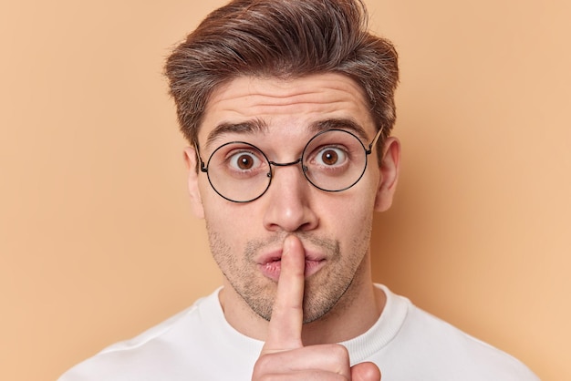 Photo close up portrait of mysterious handsome man keeps index finger over lips makes silence gesture gossips about something wears round spectacles isolated over beige background asks to keep secret safe