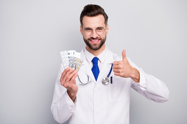 Close-up portrait of nice attractive glad cheerful doc holding in hands plates of pills excellent therapy treatment positive effect showing thumbup isolated on light white gray pastel color