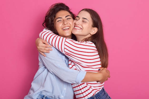 Close up portrait off positive ladies, frienads or sisters standing next to each other, having warm hug, posing with pleasant smiles, isolated over pink background. Friendship and happyness concept.