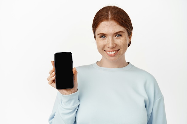 Close up portrait of redhead girl showing cellphone screen and smiling. Young woman holding mobile phone on white