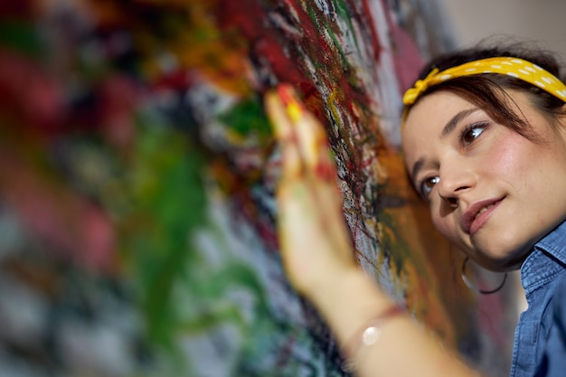 Close up portrait of relaxed young woman female painter applying paint on canvas with fingers while