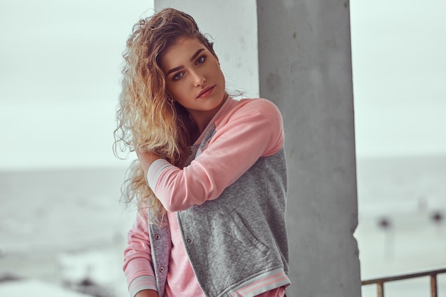 Close-up portrait of sensual young girl with curly blonde hair dressed in a pink t-shirt and jacket posing against black and white sea coast background.