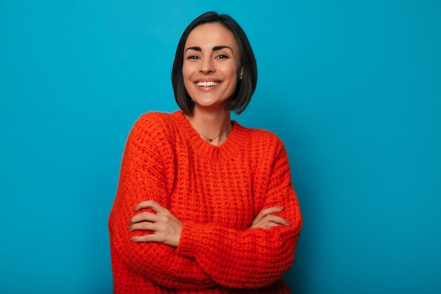 Close up portrait of smiling beautiful young brunette woman in bright color sweater is posing with crossed arms on blue background