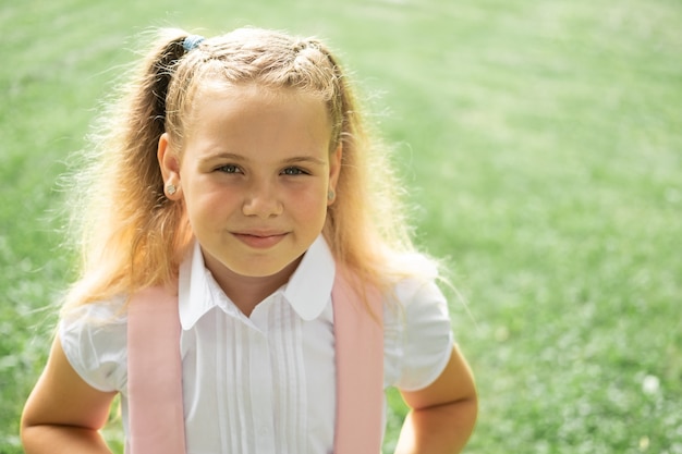 Close up portrait of smiling blonde schoolgirl in white shirt with pink backpack back to school outdoor.