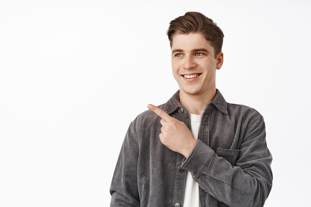 Close up portrait of smiling collegy boy, student pointing and looking left with happy face, showing banner logo, advertisement on empty space, white background