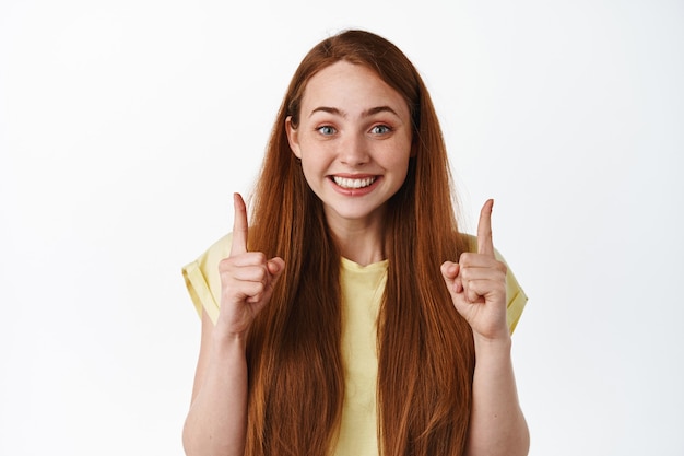 Close up portrait of smiling ginger girl with long hair, looking with excitement and pointing fingers up on top on white.