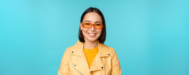 Close up portrait of smiling stylish asian woman in sunglasses looking happy at camera posing in trendy yellow jacket blue background