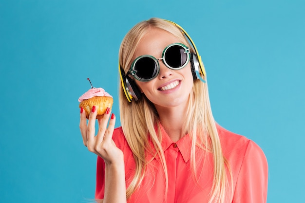 Close up portrait of a smiling young blonde woman in sunglasses listening music with headphones over blue wall