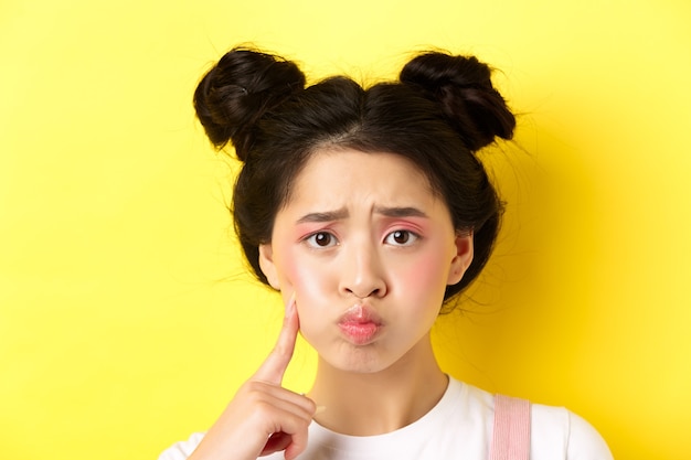 Close-up portrait of stylish asian girl with bright makeup and glowing skin, pouting and frowning, poking her cheek with sad face, standing upset on yellow