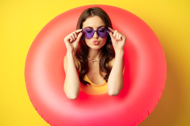 Close up portrait of stylish young woman, girl with swimming ring and summer sunglasses, ready for beach holiday, vacation, standing over yellow background