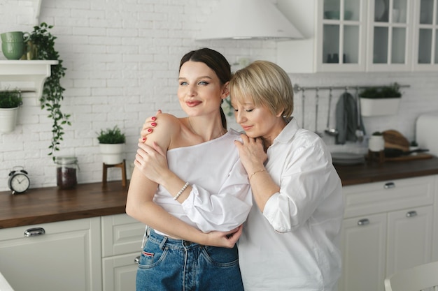 Close-up portrait of two nice attractive lovely tender cheerful women mom mommy adult daughter spending time day together in white light interior house flat apartment indoors