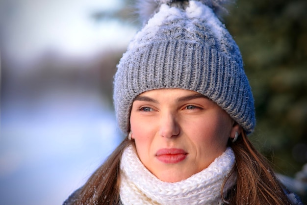 Close up portrait of unpleasant angry unhappy displeased girl, young sad frozen woman in warm clothes knitted snowy hat and scarf in snow walking in park outdoors. Cold weather, freezing person.