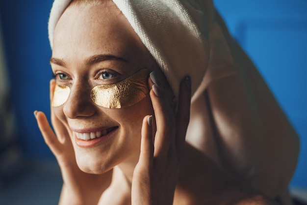 Close up portrait of a woman after shower with a white towel on her head and with patches under her ...