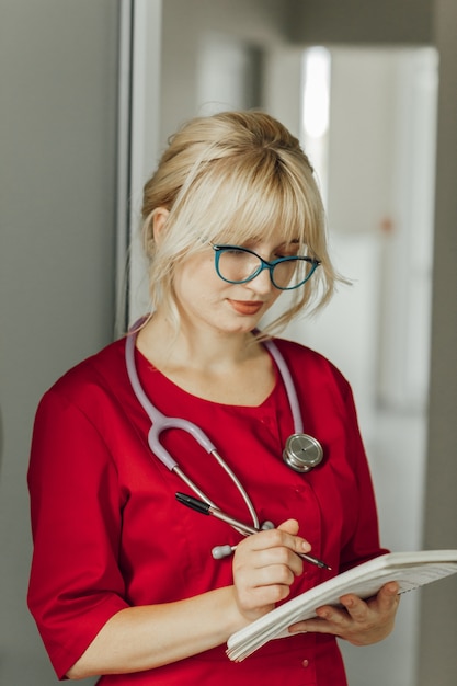 Close-up portrait of a young blonde doctor in a red uniform with a stethoscope. A doctor with glasses is in the hospital and writes something in a notebook