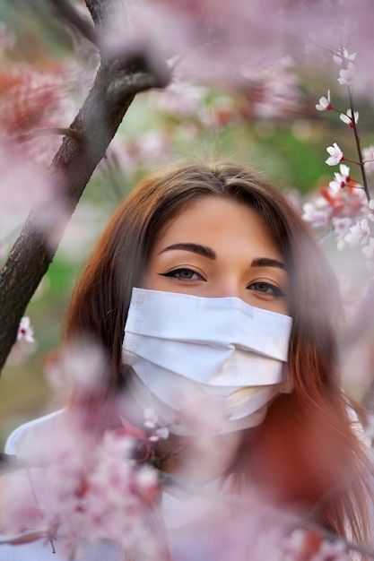 Close up portrait of young girl under a blossoming apricot tree with a mask from the coronavirus.