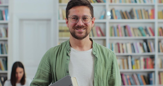 Close up portrait of young man in glasses holding digital tablet in library bookshelf background looking at the camera College education concept