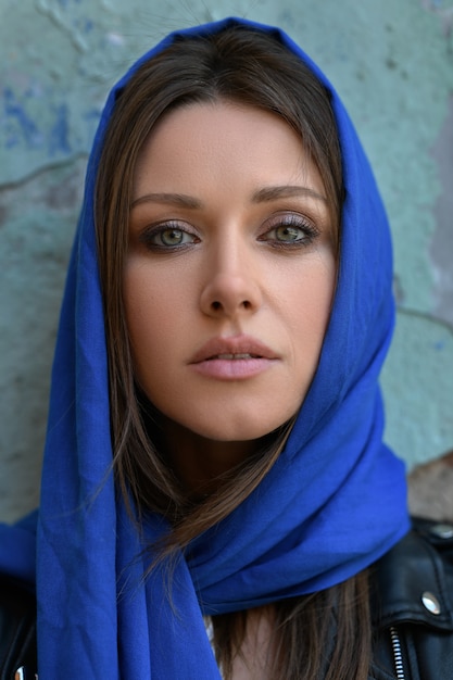 Close up portrait of young religious brunette hair green gentle eyes beautiful face adult woman or young girl with blue silk scarf covered head in black leather jacket on old brick wall background