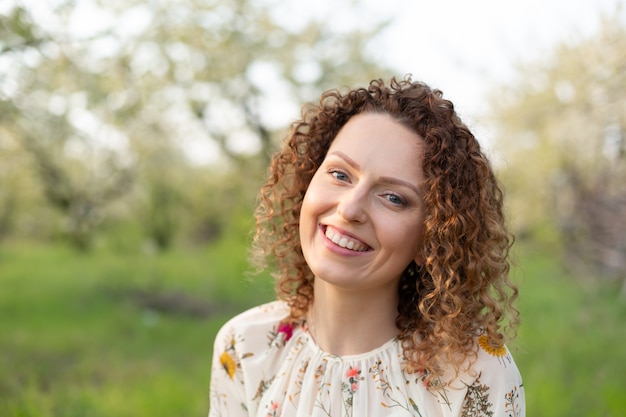 Close up portrait of young smiling attractive woman with curly hair in green flowering spring park. Pure emotions