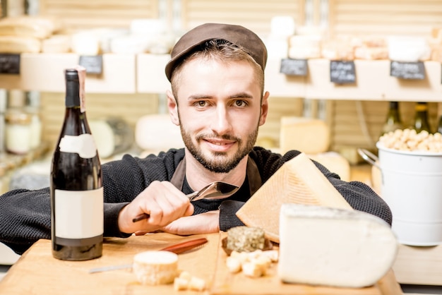 Close-up portrait of a young sommelier or cheese seller with cheese assortment and wine bottle in front of the store showcase