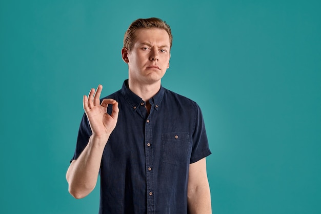 Photo close-up portrait of a young stately ginger guy in a stylish navy t-shirt showing ok sign and looking at the camera while posing on blue studio background. human facial expressions. sincere emotions c