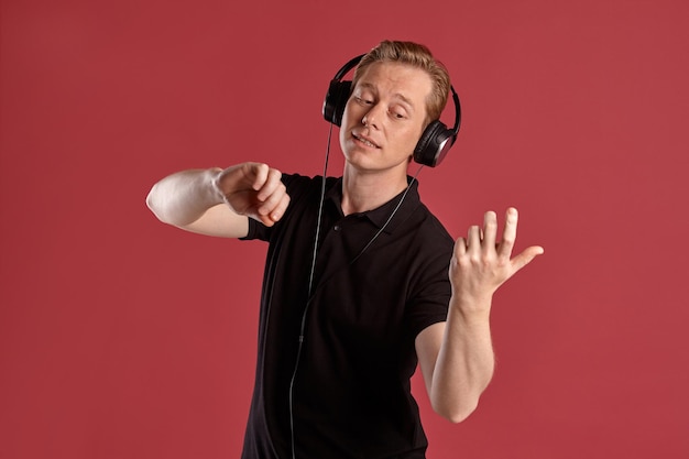 Close-up portrait of a young stylish ginger man in a stylish black t-shirt listening to the music through a headphones and pretending like he is playing the violin while posing on pink studio backgrou