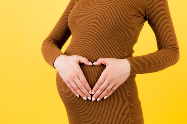 Close up of pregnant woman's belly with heart made of fingers at yellow background. Maternity concept. Love to the future baby. Copy space.