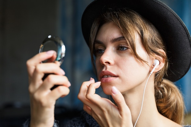 Close up of a pretty young woman in hat fixing her make up while looking at the mirror indoors